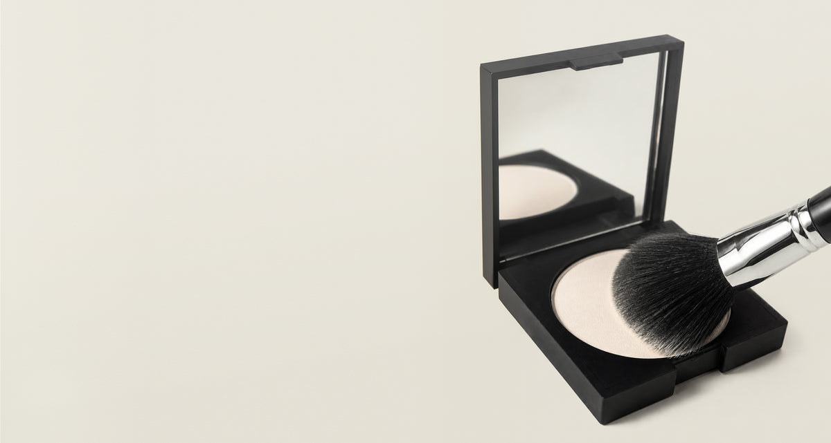 An open black square makeup compact with a mirror and a large black-and-silver makeup brush resting on a pressed powder, shown on a plain light beige background.